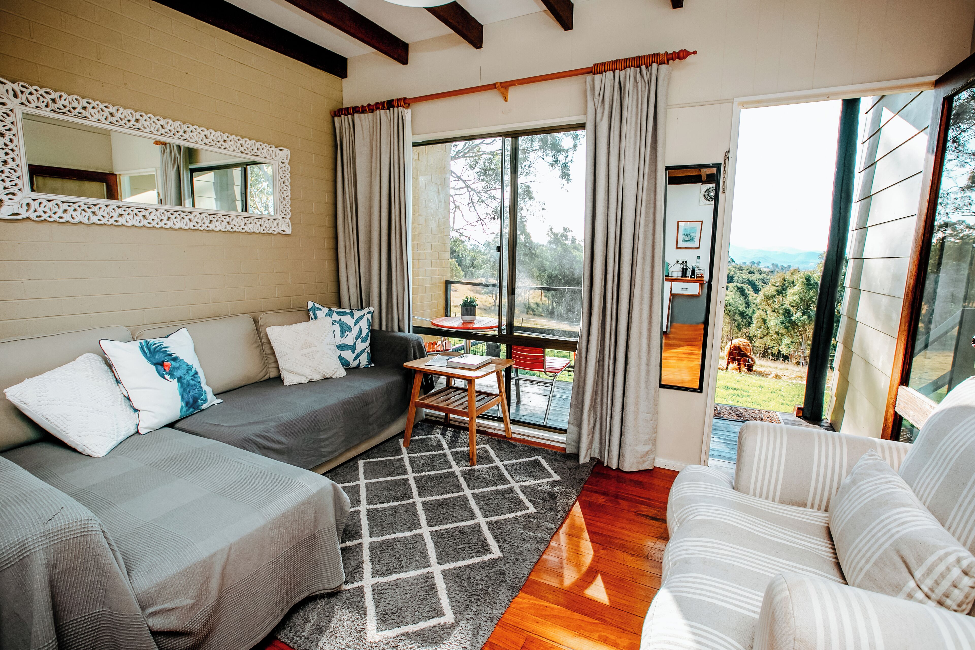 A cozy living room with a grey sofa, a striped sofa, and a small wooden table. There is a large window with curtains, a mirror on the wall, and a view of the outdoors.