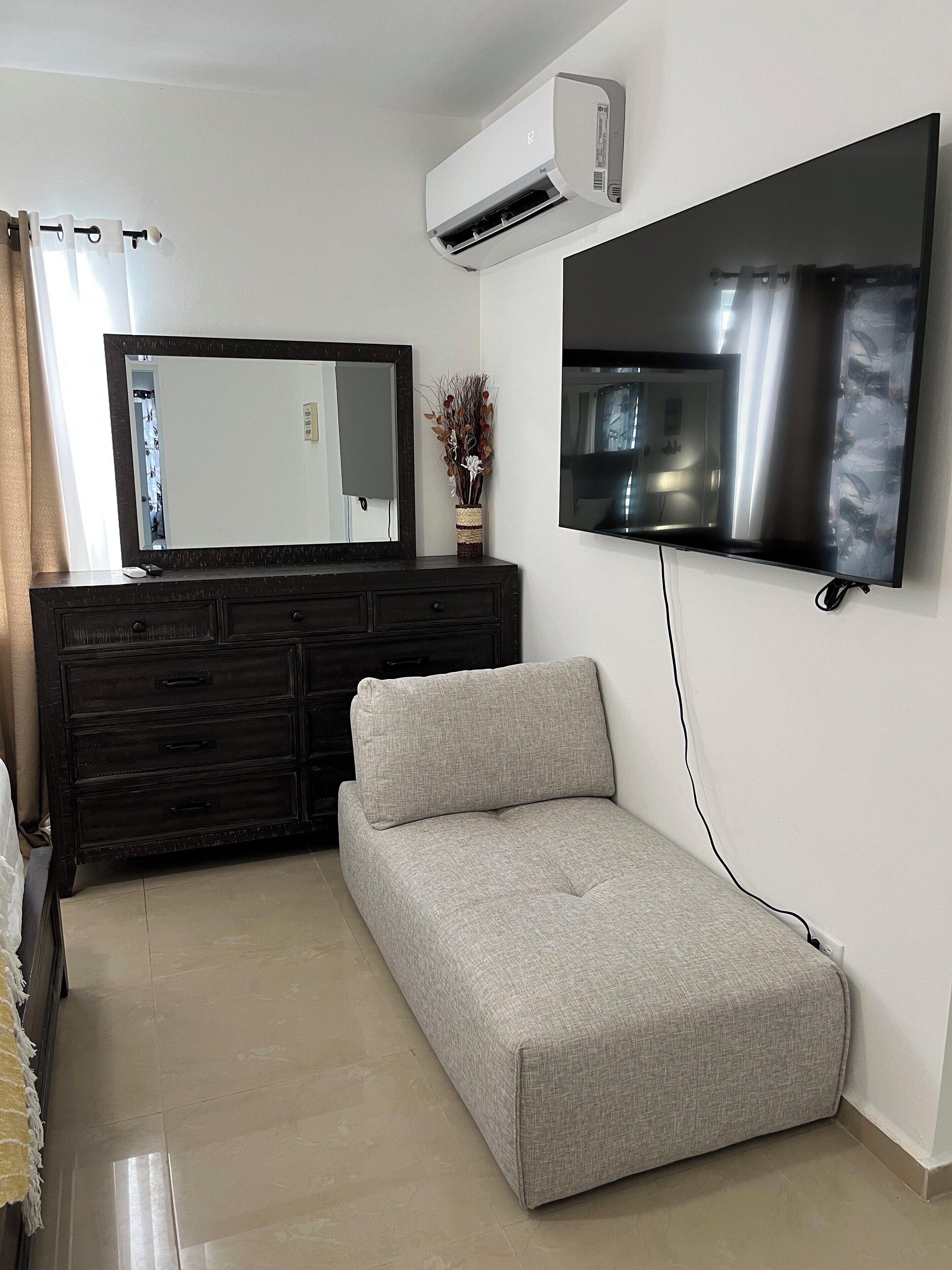 A modern living room with a dark wooden dresser, a grey sofa, a flat-screen TV mounted on the wall, and an air conditioning unit above the dresser.
