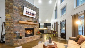 A spacious living area with a stone fireplace, a kitchen in the background, and a large window with curtains. - Cozy Nights Await at Ember & Stone– 3 Fireplaces, Hot Tub & Tucked Away Vibes (Broken Bow)