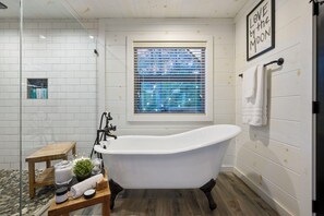 A bathroom with a freestanding bathtub, a small wooden stool, a window with blinds, and a wall-mounted towel rack.