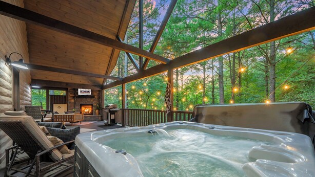 A hot tub in a wooden deck area with a stone fireplace and outdoor seating. - Cozy Nights Await at Ember & Stone– 3 Fireplaces, Hot Tub & Tucked Away Vibes (Broken Bow)