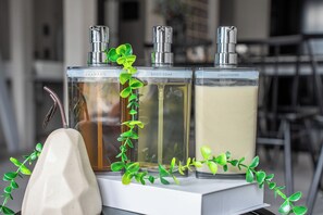 Trois bouteilles de shampoing, de savon pour le corps et d’après-shampoing, posées sur une surface blanche, avec des plantes vertes à proximité.