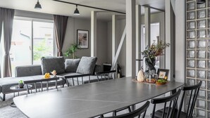 A modern dining area with a large table, grey sofas, and a staircase leading to another level. - SNOWGARDEN YIG STAY (Furano)