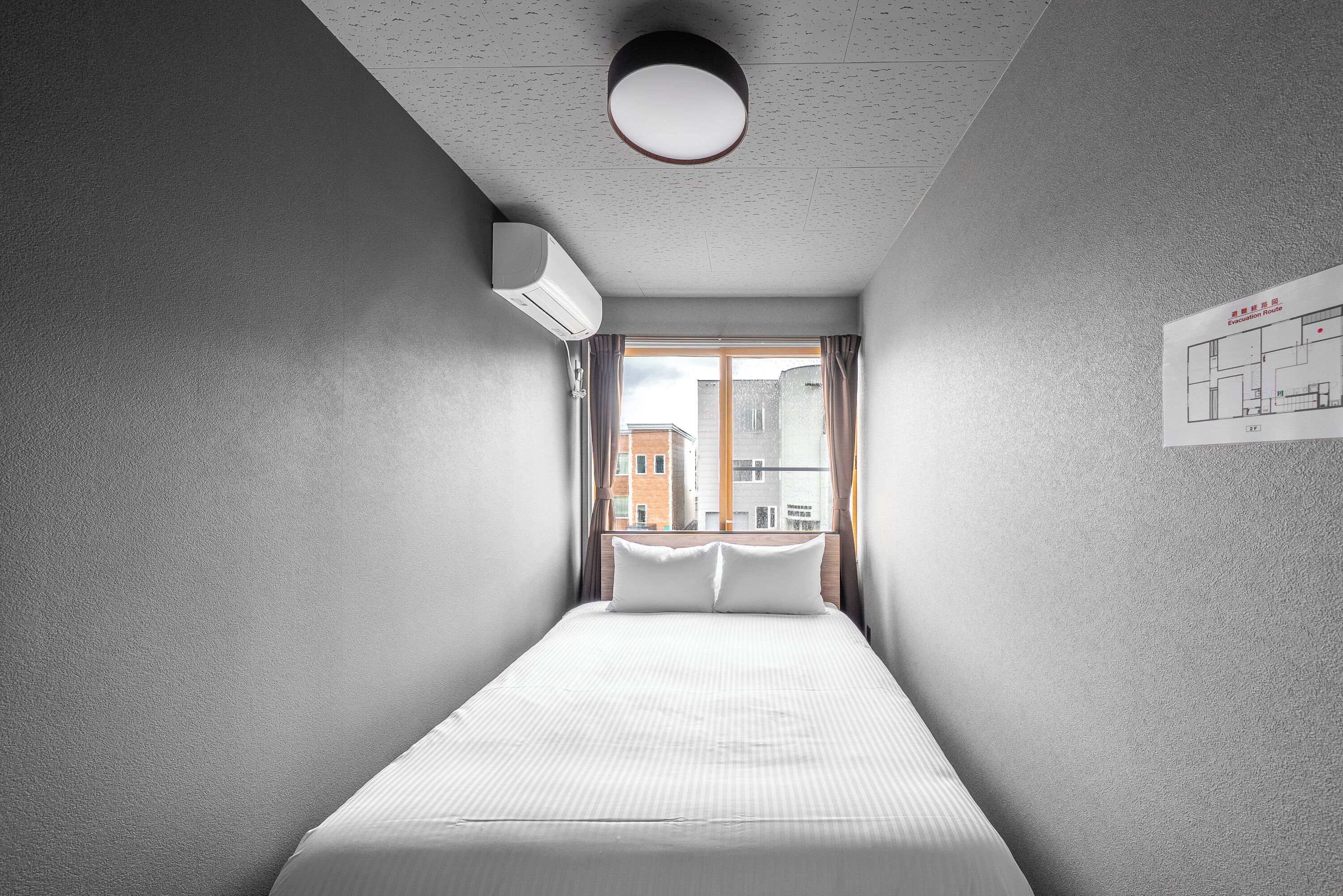 A narrow hotel room with a single bed, a window with curtains, a ceiling light, and a wall-mounted air conditioning unit.