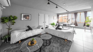 A modern, well-lit room with two beds, a dining table, and a large window providing a view of the city. - WHITEGARDEN YIG STAY (Furano)