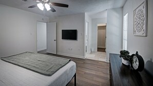 A bedroom with a bed, a ceiling fan, a TV mounted on the wall, and a small table with a clock and a plant. - The Rez Retreat - King Beds (Brandon)