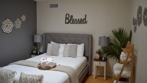 Ein ordentlich eingerichtetes Schlafzimmer mit einem großen Bett, zwei Nachttischen mit Lampen, einem an der Wand hängenden Kunstwerk mit der Aufschrift "Blessed" und einer kleinen Pflanze auf einem Tisch.