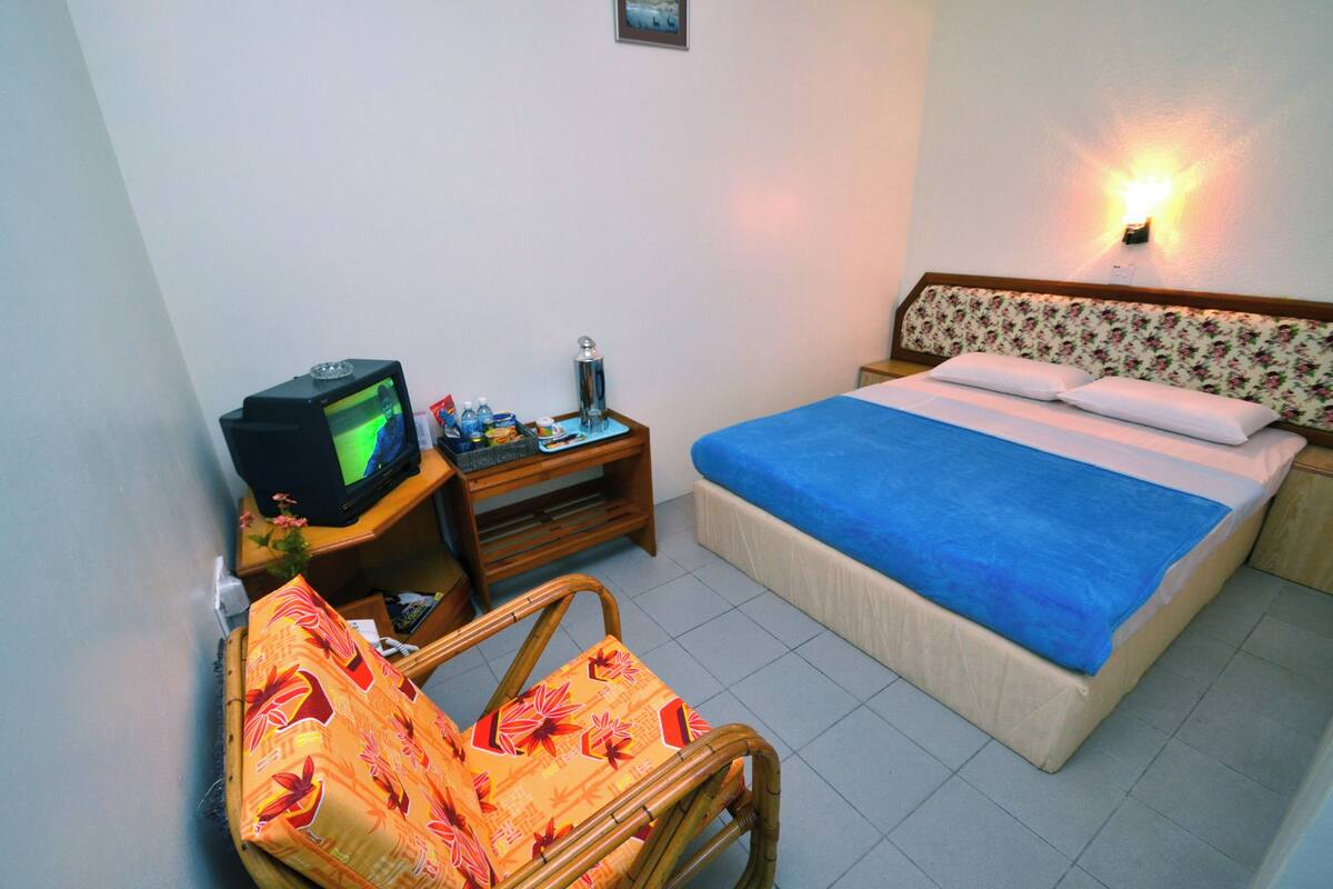A bedroom with a bed, a television on a wooden stand, a chair, and a wall-mounted light.