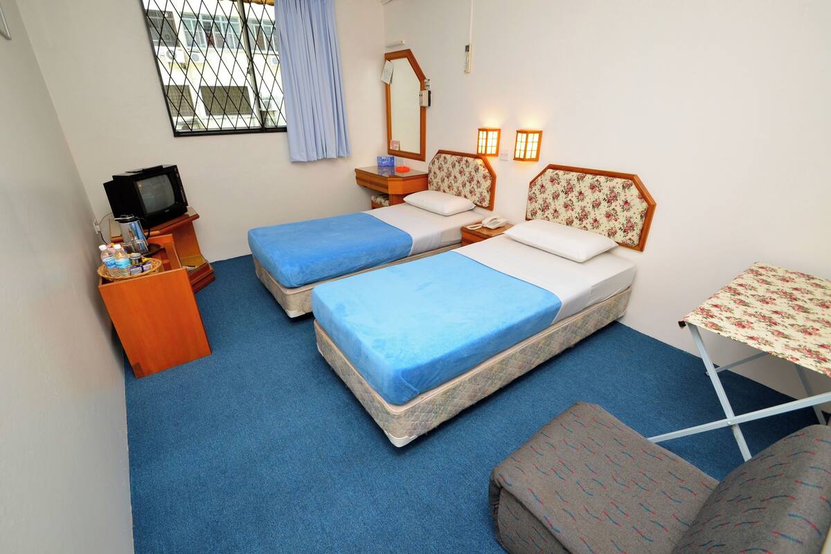 A small, simple room with two beds, a small table, a TV, and a window with curtains.