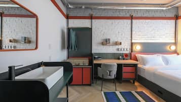A compact hotel room with a bed, a desk, a chair, a small table, a mirror, and a brick-patterned wall.