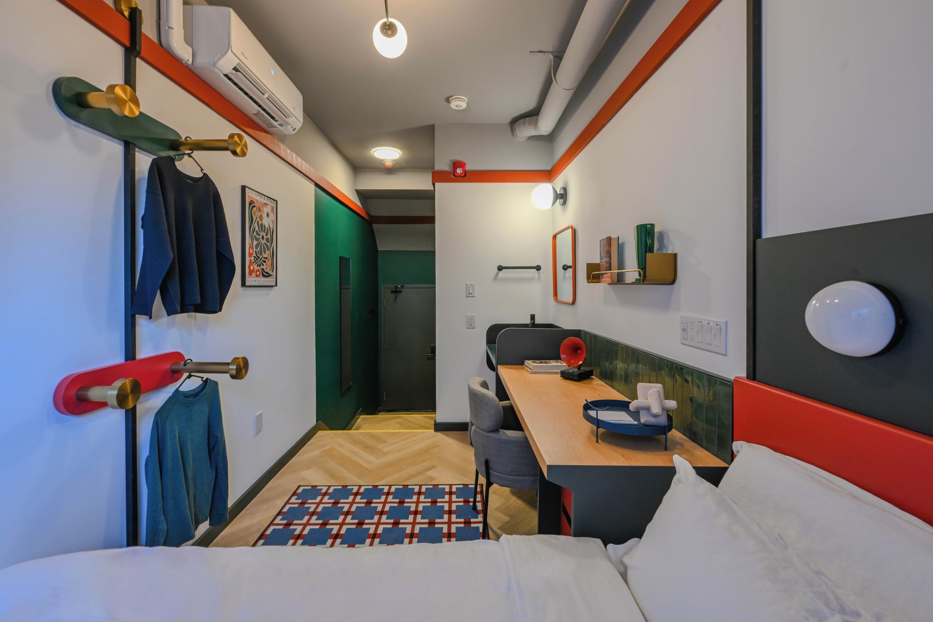 A compact hotel room with a bed, a desk, a chair, a small table, a mirror, and a wall-mounted heater.