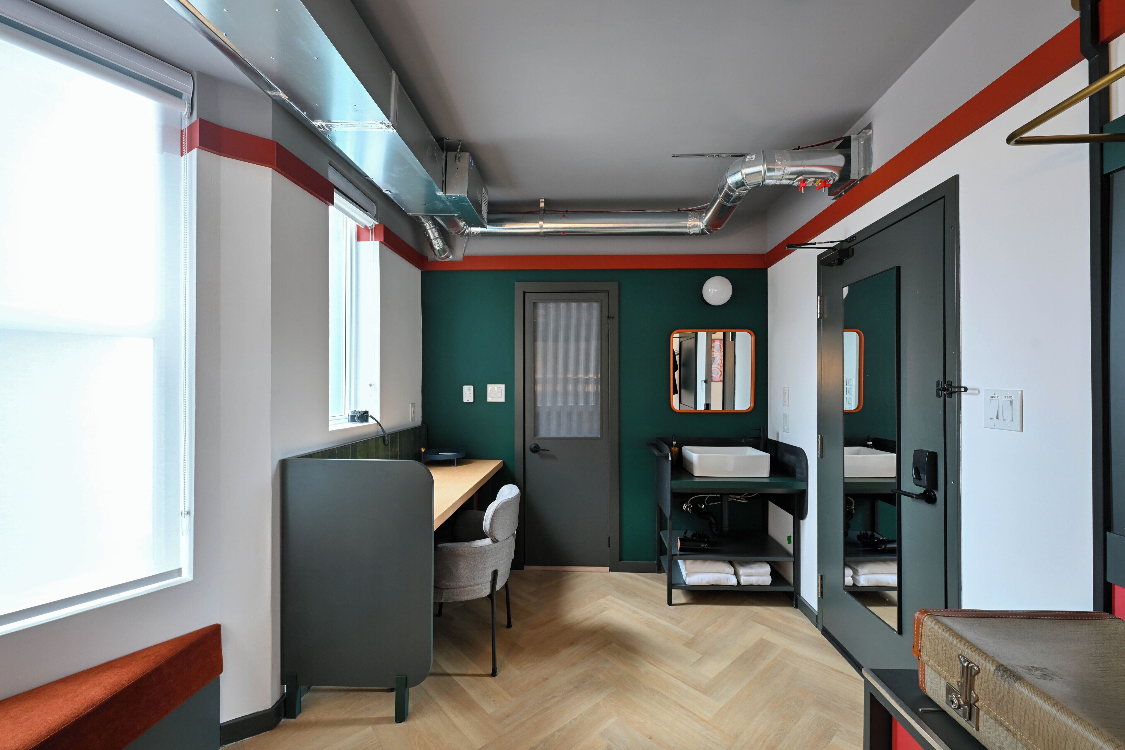 A compact room with a desk, chair, and a bed with storage. There is a window providing natural light and a door leading to another room.