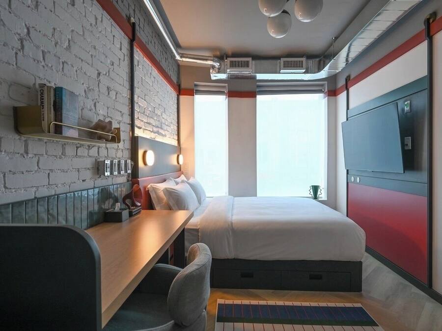 Habitación de hotel pequeña con pared de ladrillo, cama, escritorio con silla, televisor y ventana con vistas.