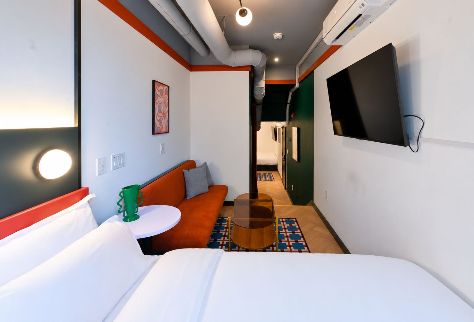 A hotel room with a bed, a sofa, a small table, a TV mounted on the wall, and a hallway leading to a bathroom.