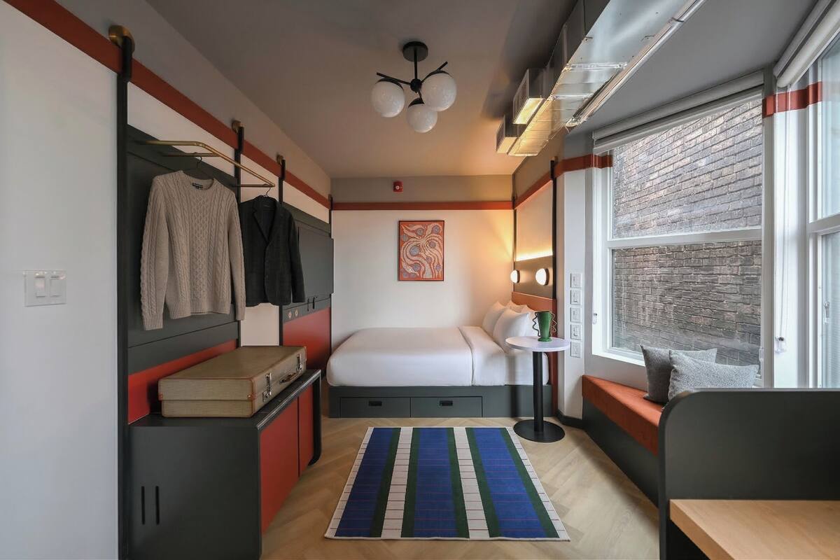 A compact hotel room with a bed, a small table, a chair, a wardrobe, a suitcase, a window with a view, and a ceiling fan.
