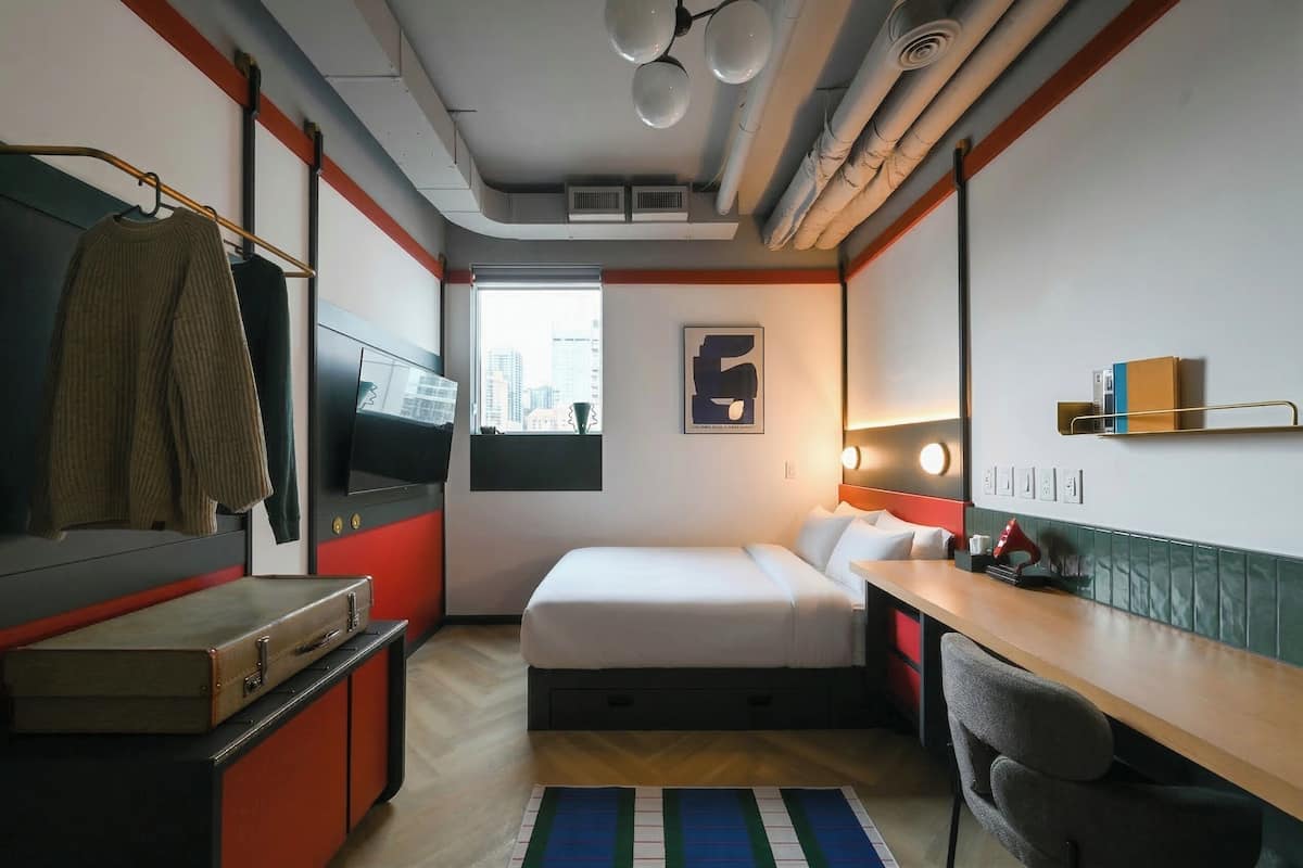 A compact hotel room with a bed, desk, chair, and a view of the city through the window.