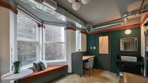 A modern room with a large window, a desk, a chair, a small table with a green vase, and a mirror. - Nomas Hotel (Toronto)