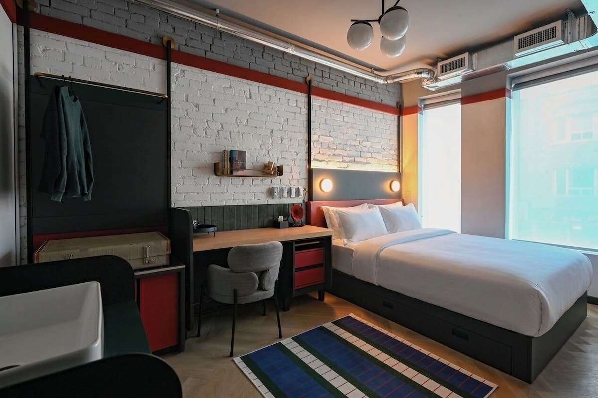 A modern hotel room with a brick wall, a bed, a desk, and a chair.