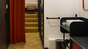 A hallway leading to a bedroom with a bed, a bathroom with a sink and mirror, and a trash bin. - Nomas Hotel (Toronto)