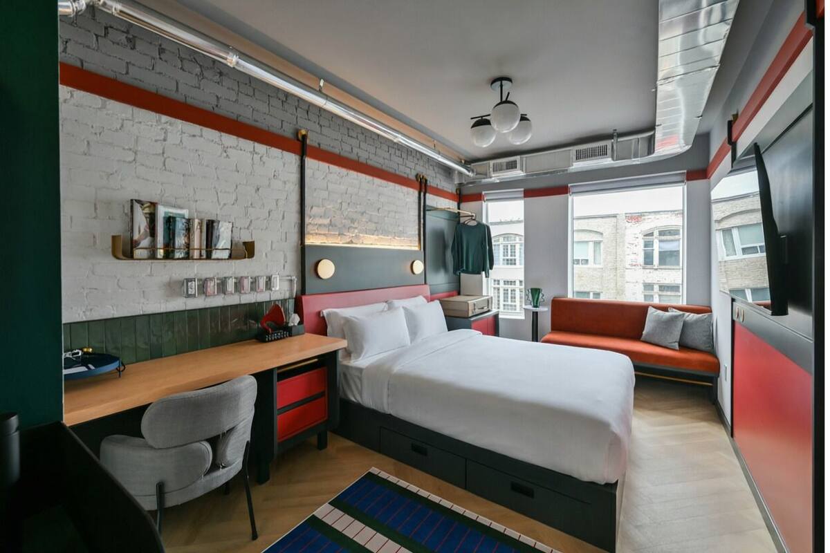 A modern hotel room with a brick wall, a bed, a desk, a chair, a sofa, and a TV.