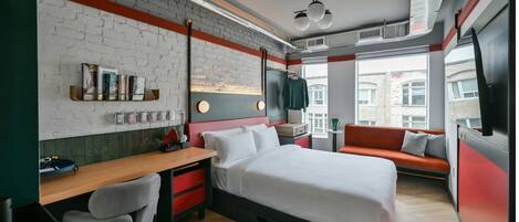 A modern hotel room with a brick wall, a bed, a desk, a chair, a sofa, and a TV.