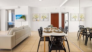A modern living room with a sofa, dining table, and chairs. There are framed pictures on the wall and a view of the outdoors through a window. - Chevron Renaissance Ocean View (Surfers Paradise)
