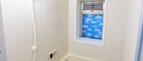 Ein kleines, weißes Badezimmer mit Dusche, Toilette und einem Fenster mit Blick auf einen blauen Himmel und Wolken.