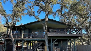 A two-story wooden cabin with a balcony, surrounded by trees and a grassy yard.