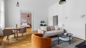 Un salon moderne et bien éclairé, avec un canapé, une table basse et des fauteuils. On aperçoit à l’arrière-plan un coin repas avec une table et des chaises.