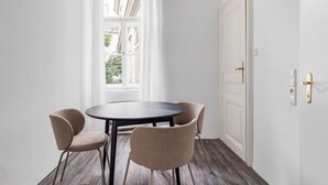 Une pièce avec une table ronde et quatre chaises, une porte blanche avec une poignée en laiton et une fenêtre ornée de rideaux blancs.