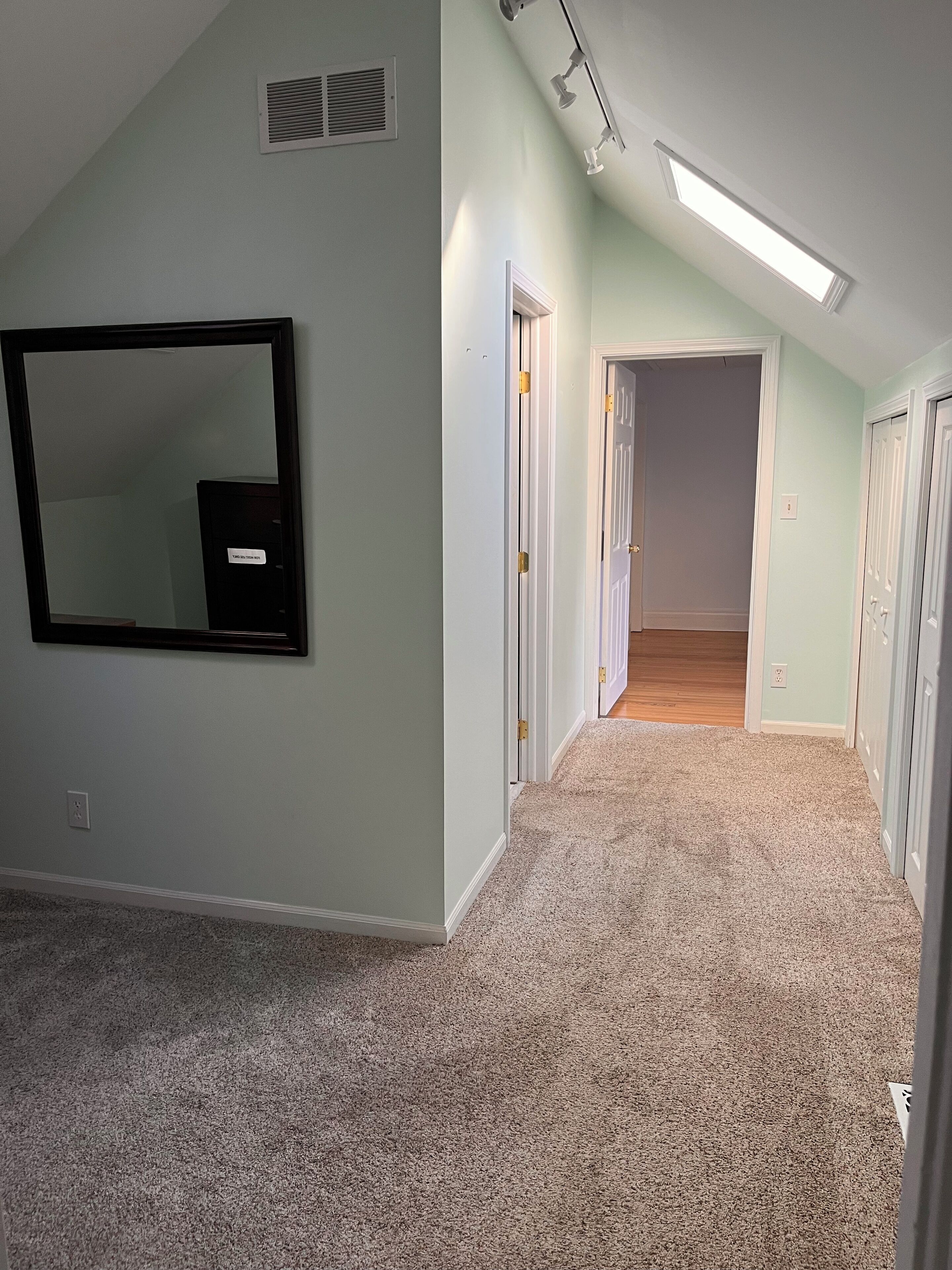 A hallway with carpeted flooring, a ceiling vent, a mounted mirror, and a door leading to another room.