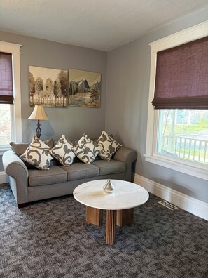 A living room with a grey sofa, patterned cushions, a round coffee table, and two wall-mounted paintings.