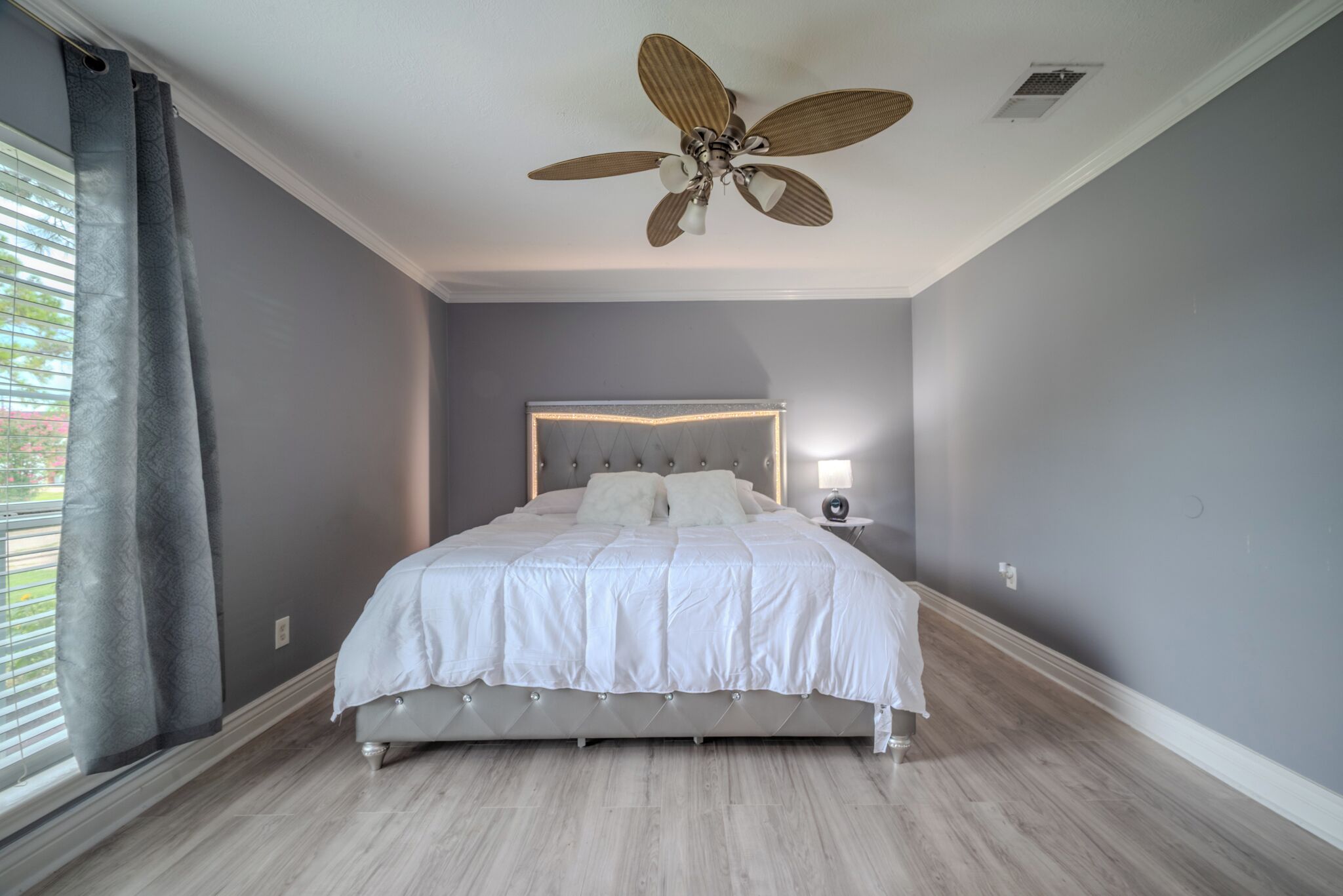 A bedroom with a bed, a ceiling fan, a bedside table with a lamp, and a window with curtains.