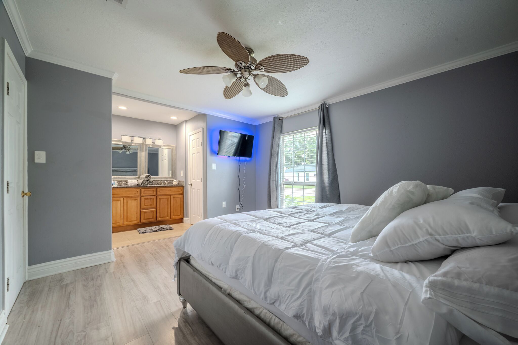 A bedroom with a large bed, a ceiling fan, a TV mounted on the wall, and a bathroom visible in the background.