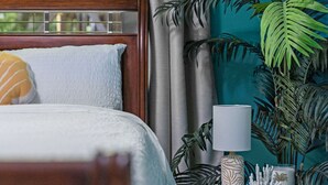 A bedroom with a bed, curtains, a window with a view of a playground, a potted plant, and a bedside table with a lamp and decorative items. - The Quirky Casita in League City (League City)