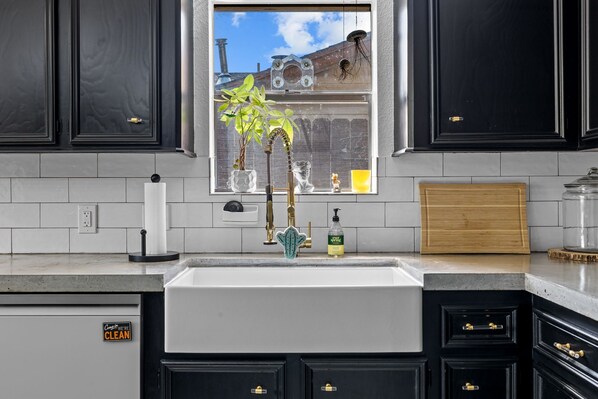 Una cocina con armarios oscuros, un lavamanos blanco, una ventana con vista a un muro de ladrillo y una tabla de cortar de madera sobre la encimera.