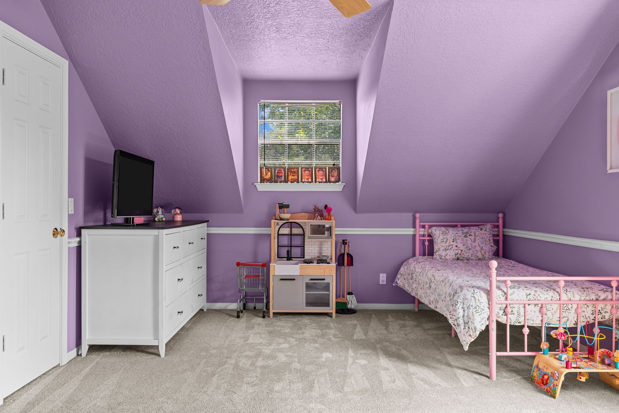Une chambre d’enfant avec des murs violets, une commode blanche, un lit rose et une fenêtre avec des stores.