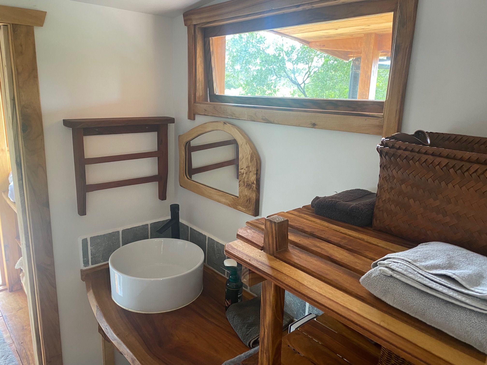 Ein kleines Badezimmer mit einem Holzbadezimmerhocker, einem runden Waschbecken, einem Handtuch und einem Fenster mit Blick auf Grünflächen.