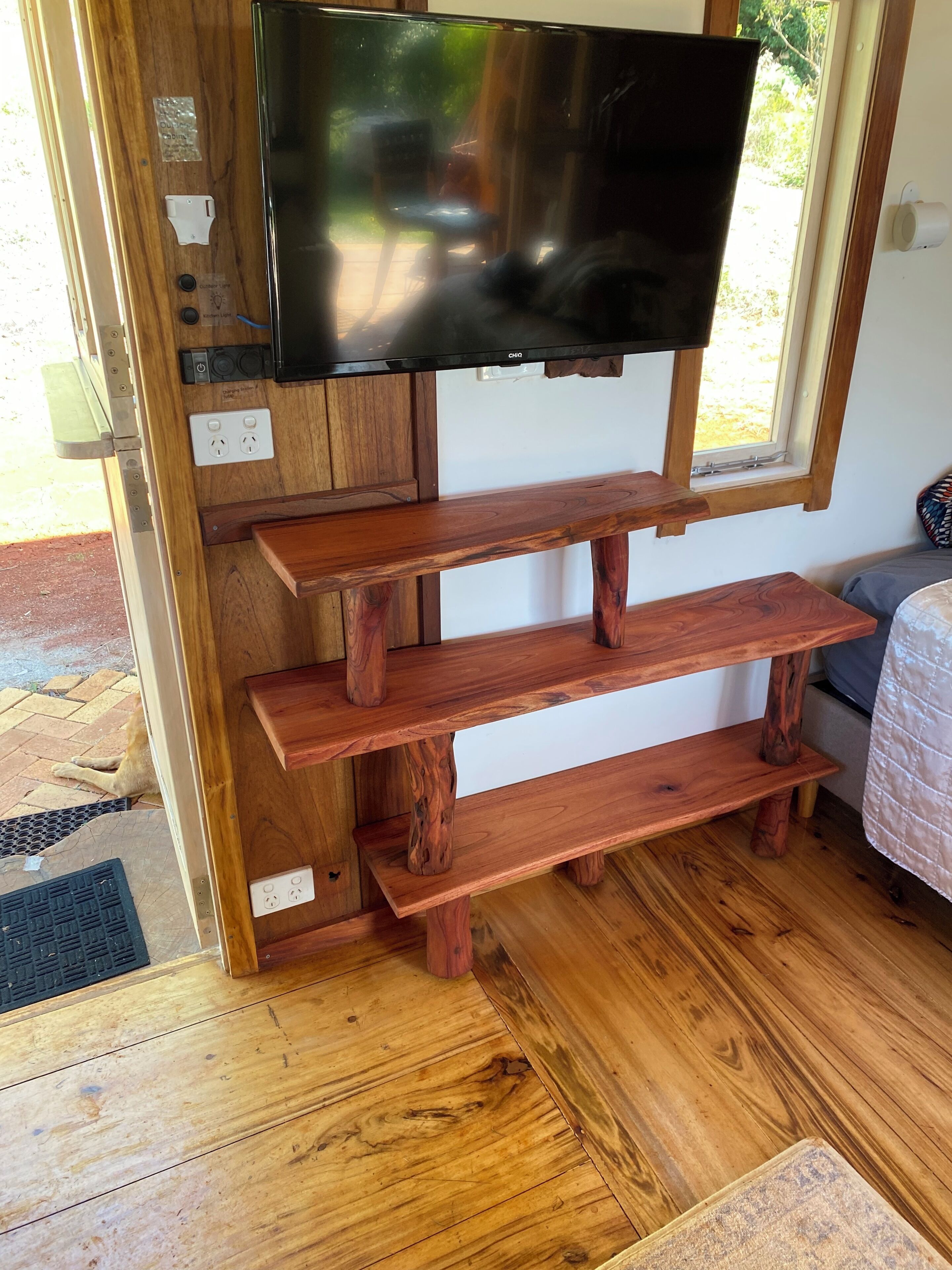 A wooden TV stand with a flat-screen TV mounted above it, a window with a view of greenery, and a power outlet on the wall.
