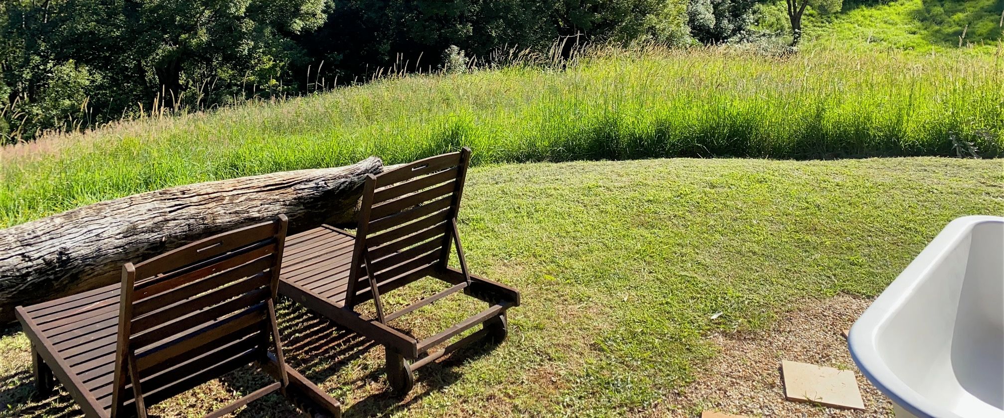 A wooden bench with a log on it, positioned on a grassy area with a white structure nearby.