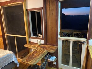 A room with a wooden table, a bed, and a view of the mountains through a door.