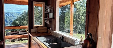Interior de una cabaña de madera con una zona de cocina, armarios de madera, un lavamanos y un ventanal con vistas a un paisaje boscoso.