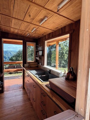 L’intérieur d’un chalet en bois comprend un coin cuisine, des placards en bois, un évier et une fenêtre donnant sur un paysage boisé.