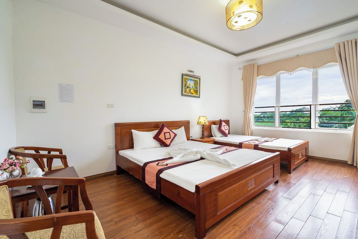 A hotel room with two wooden beds, a dining table with chairs, a small table with a vase of flowers, and a window with curtains.