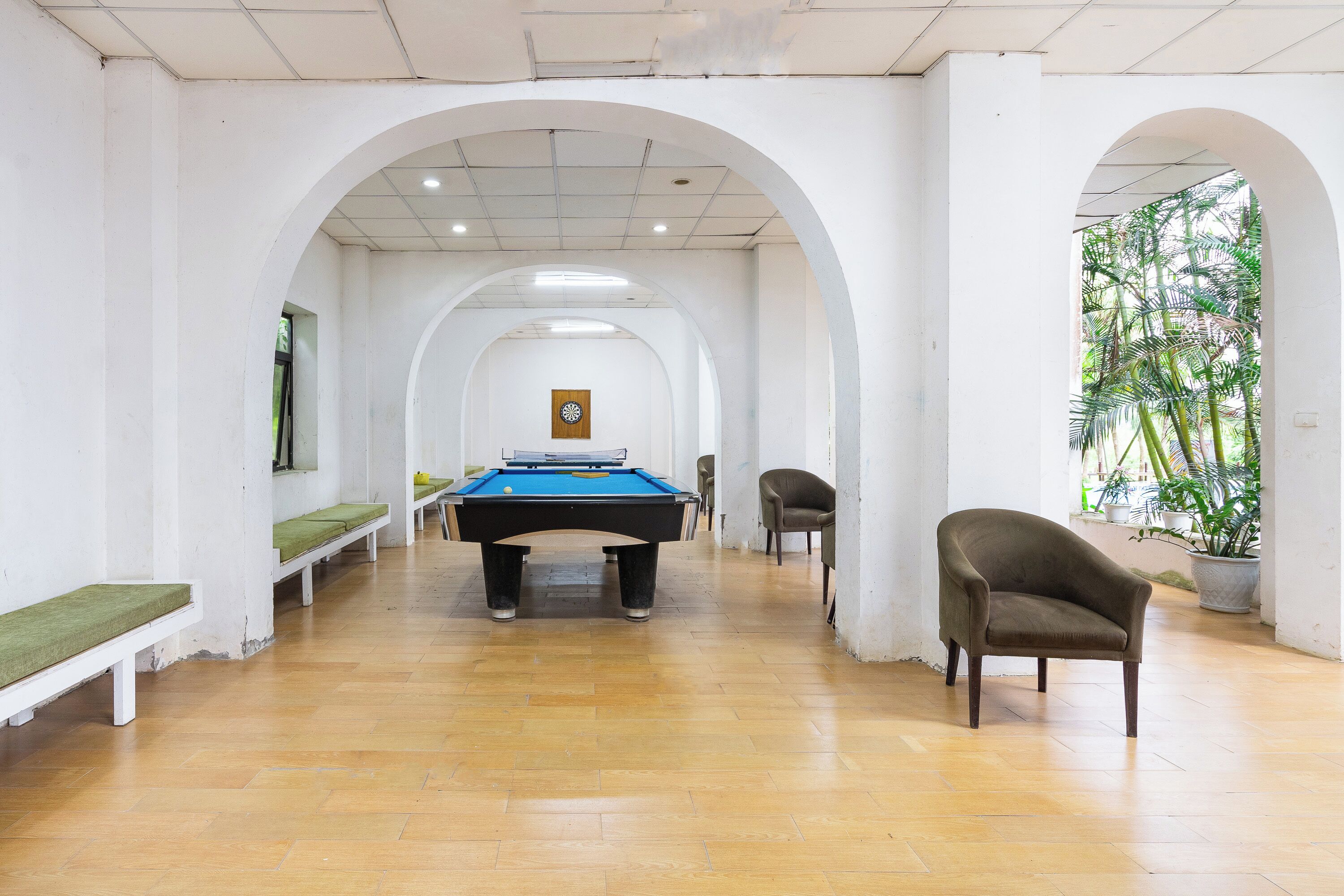 A pool table in a hallway with benches and a chair, white walls, and a view of greenery outside.