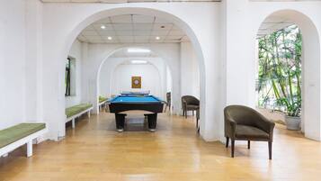 A pool table in a hallway with benches and a chair, white walls, and a view of greenery outside.