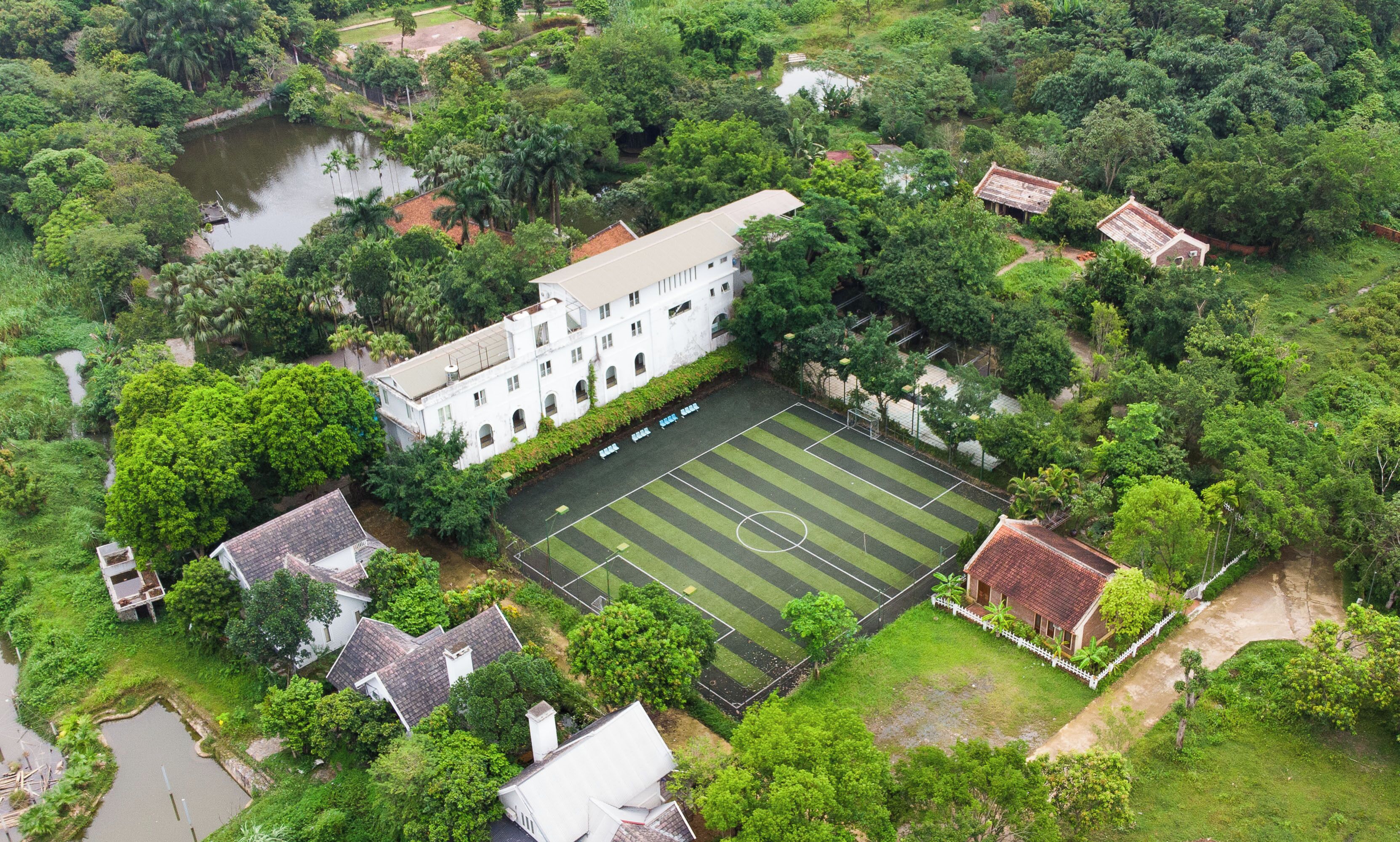Vista aérea de un edificio grande rodeado de exuberante vegetación, con un campo deportivo al frente.