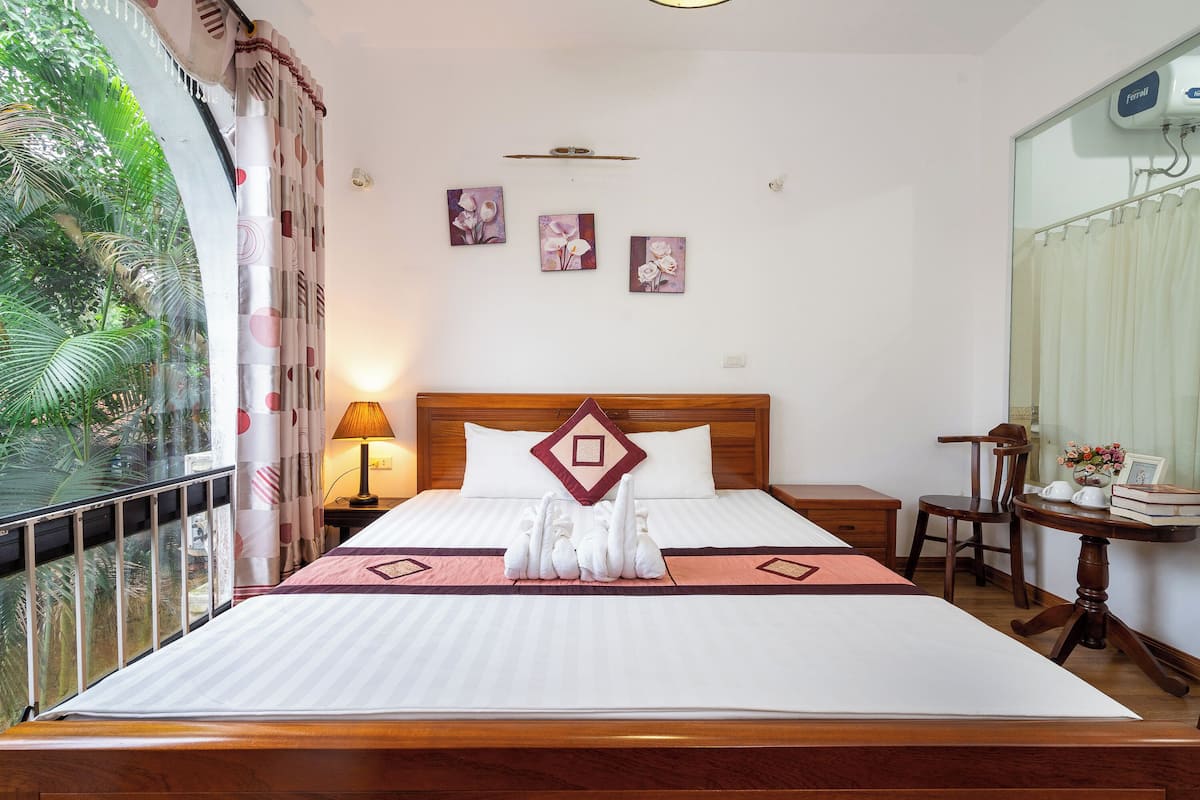 Una habitación de hotel con una cama grande, un cabecero de madera, una mesita de noche con lámpara, una pequeña mesa con libros y un jarrón, y una ventana con cortinas.