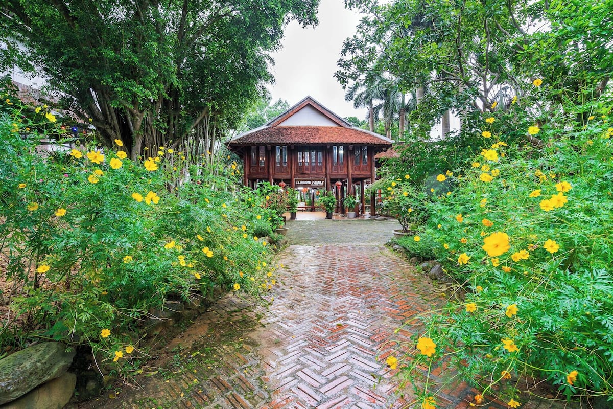 Una casa de madera tradicional con un sendero de ladrillo en espiga que lleva a la entrada, rodeada de abundante follaje y flores amarillas.