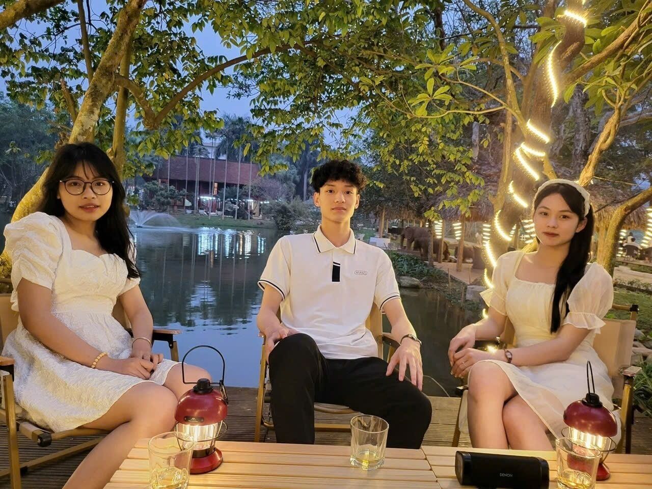 Trois personnes sont assises à une table, devant des boissons, et sont entourées d’arbres et d’une étendue d’eau.